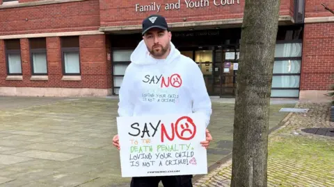 Simon Cobb Simon Cobb is standing outside a Family  and Youth Court with a poster which says "say no to the death penalty; loving your child is not a crime". He wears a white hoodie and black cap