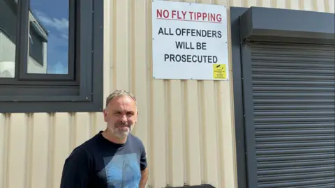 McClelland stands in a blue t-shirt in front of a sign on a tin building. The sign reads: 'NO FLY TIPPING ALL OFFENDERS WILL BE PROSECUTED'.