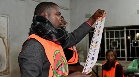 An election official wearing an orange tabard and scarf holds up a ballot paper for inspection.