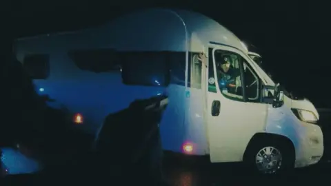 Channel 4 A white camper van is on a road and the drivers door is open. Stephen Alderton is sat on the drivers seat with his hands up. 