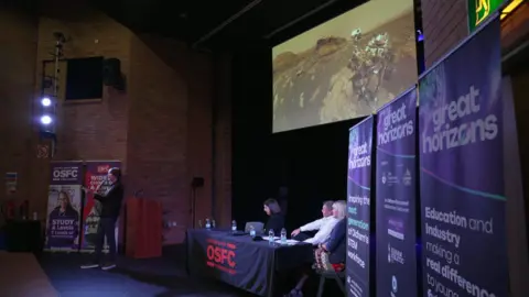 Professor Brian Cox stands in front of a screen with a picture of a robot on the planet Mars. Three people sit at a table behind him. On the right hand side of the picture are purple boards advertising the Great Horizons events