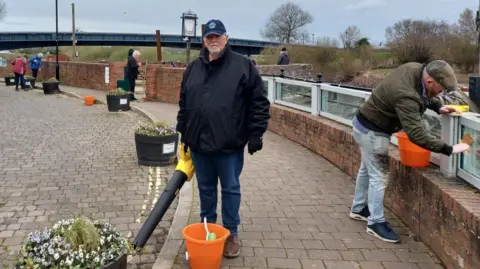 Steve Dunwell A man wearing a blue hat, with short grey hair and a grey beard, holds a leaf blower. He is wearing a navy blue coat and blue jeans. Members of the community clean in the background.
