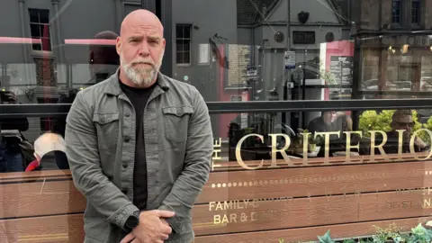 Steve stands in front of his pub, arms folded. He wears a green jacket and has a beard. 