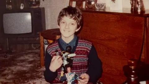 Family Mark Billington, 15, in a living room in the 1980s, holding an Action Man figure boy and other toys