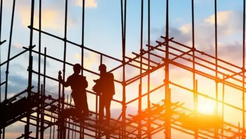 Two builders stand on scaffolding as the sun sets behind them on a construction site.