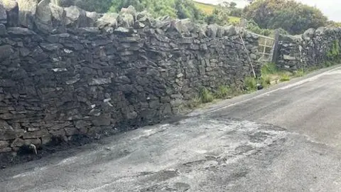 A fire-damaged section of tarmac, next to a stone wall. A tall brush has been propped up next to a gate.