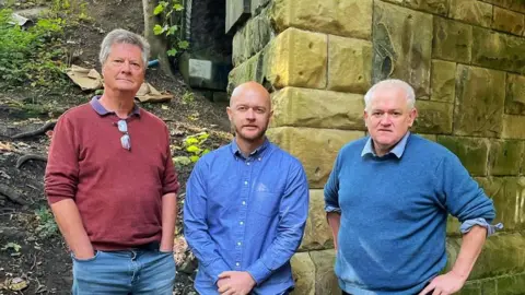 SELRAP campaigners Chris Oakley, left, and Peter Bryson, right, with Pendle & Clitheroe MP Jonathan Hinder, all wearing casual clothing and standing by a stone railway tunnel next to a steep embankment