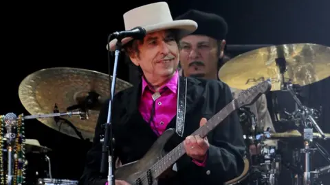 A man in a pink shirt and black jacket, wearing a hat, and holding a guitar. behind him is a man playing a drumkit.