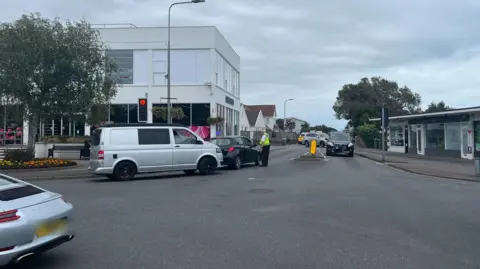 A police officer talks to a driver in a black car as colleagues further up the road deal with a car crash. Other vehicles including a silver Porsche and silver van are on the road. An M&S Food shop in in the background.