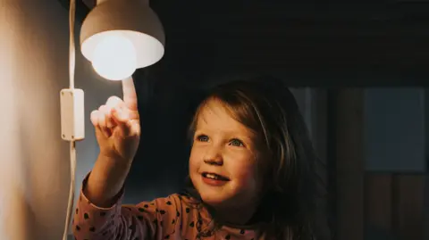 Getty Images A small girl pointing at a lamp which is switched on. She has blue eyes and dark blonde hair and is smiling at the lamp. 