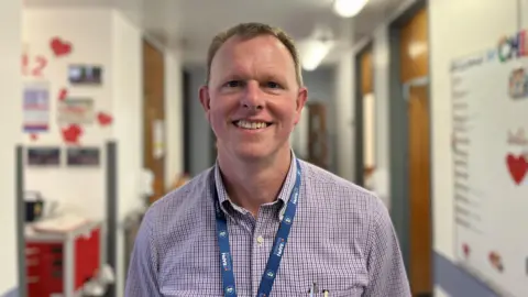 A man with short light brown hair smiles into the camera. He is wearing a blue lanyard and a checkered purple shirt. 
