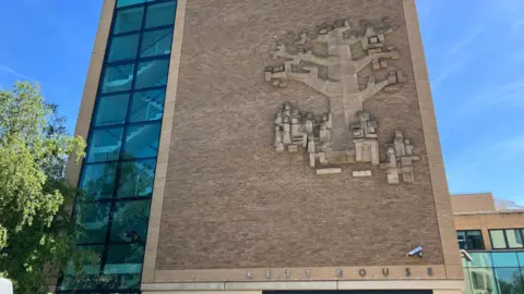 The corner wall of Kett House in Cambridge. The left third is glazed and shows interior stairs. The right two thirds is brick and on its side is an abstracted 1960s design representing an oak tree and people underneath it.