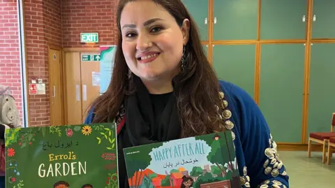 Uzra Taqwa smiles at the camera while holding two childrens storybooks which are dual language