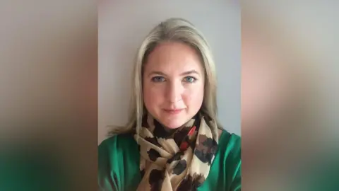 Handout A blonde woman wearing a green shirt and a brown and black patterned scarf. 