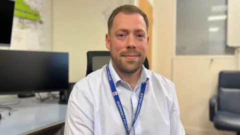Thomas Lowe pictured in a white button down shirt. He has short brown hair and a beard. He is wearing a blue Guernsey Police lanyard.