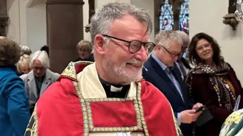 BBC Nigel Godfrey with red and cream ceremonial robes over in his dog collar and black top. He has grey hair and wears eye glasses.