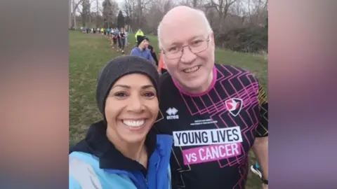 Mark Halliwell Dame Kelly Holmes and Mark Halliwell, a BBC Radio Gloucestershire commentator with grey hair, glasses and a black and pink Cheltenham Town Young Lives vs Cancer top on, smile as they take a selfie at Cheltenham parkrun. Other runners can be seen running along the path made in the grass behind them. Dame Kelly is wearing a blue hi-vis tabard, a blue coat and a black beanie.