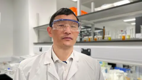 Professor Zongbo Shi is the lead investigator of the study. Here he is in a laboratory at the University of Birmingham. He has plastic goggles across his eyes and has short dark hair. He wears a white lab coat over a white shirt. Behind him are shelves with various bottles with coloured liquid in them