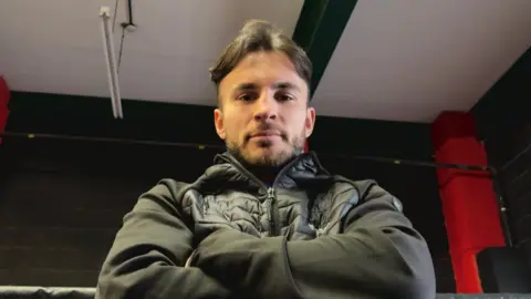 Owen Cooper, a man in a black coat has his arms crossed and is looking directly at the camera