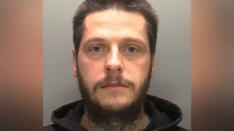 Police mug shot of a man with spikey dark hair and a beard, He is wearing a dark-coloured hoodie and has a tattoo on his neck.