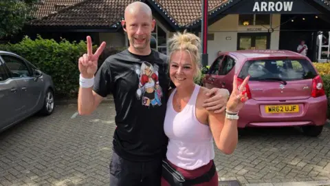 Giles and Katy stood side by side making peace signs with their hands and smiling at the camera. They are stood outside in a pub car park in running gear.