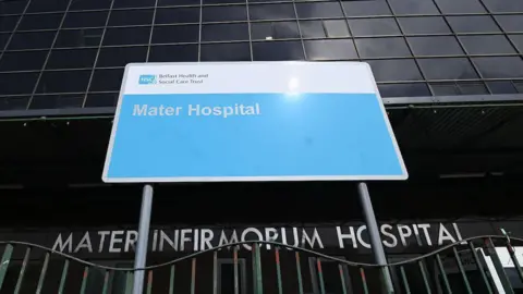 Front of the Mater Hospital. Green wavy railings are in the foreground and a sign reading 'Mater Infirmorium Hospital' in silver letters is behind. Another blue sign with white text says 'Mater Hospital'. Dark windows are above. Photo taken from below.