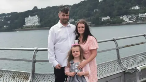 Melonie Bray Melonie Bray wearing a pink dress, with her arm around partner Ricky and daughter Sophie. Ricky is wearing a white shirt and black trousers. Sophie is wearing a silver dress with a pink cardigan and bow in her hair.