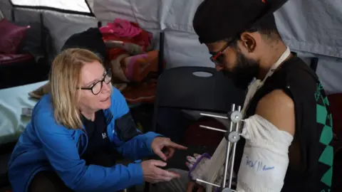 UK-Med A woman with blonde hair and glasses, wearing a blue jacket, talks to a man in a black baseball cap and black top. His left arm and hand are bandaged and in a sling, with the woman pointing at his hand.