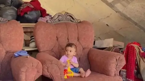 Tahani Shehada An image of Tahani Shehada's eight-month-old baby sitting on an armchair while holding a toy shovel.