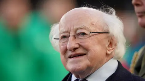 Michael D Higgins is bald on top with wispy white hair at the sides and black-framed glasses. He is wearing a dark suit