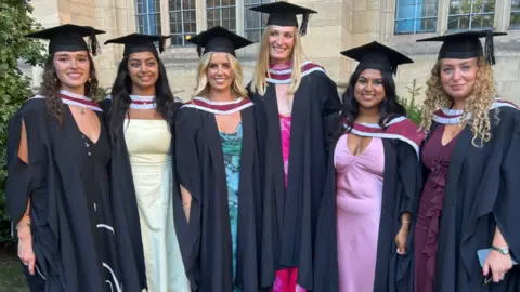Claudia Slater Six female graduates stand side by side in gowns and mortar boards. They are smiling at the camera and are standing in front of the Wills Memorial Building, a tall neo-Gothic tower in Bristol.