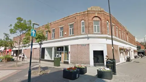 A brick-built, two-storey building in a town centre. There are pedestrians walking down the side of the building and there are raised flower beds in black containers in front of the building.