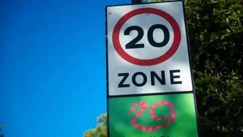 Road sign showing a 20 mph speed limit zone with snake graphic at the approach to a residential area in Fulham