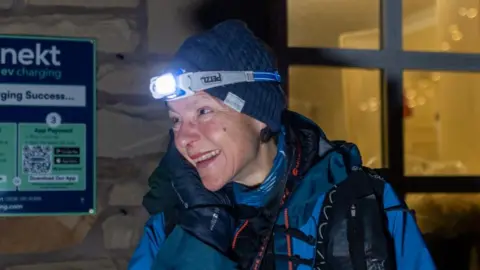 Adam Jacobs Anna Troup at the finish line at Kirk Yetholm. She is in front of a building and she is looking to the side and smiling. She is wearing a navy woolly hat and a headtorch, which is lit. She also wears a bright blue outdoor jacket.