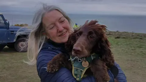A cocker spaniel in the arms of a women with long grey hair on a grassy cliff top