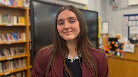 Rachel has long brown hair and is smiling at the camera. She is standing in the school library in front of a large screen. She is wearing a maroon school blazer, a striped shirt and a navy school jumper.