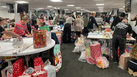 An office building floor with lots of people at tables wrapping presents. The floor is covered in boxes of wrapped presents.