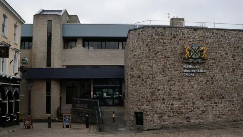 Ian S/Geograph The front entrance of Plymouth Magistrates' Court on a cloudy day. A large sign for the court is on a brick wall. A pub is to the left of the court building.
