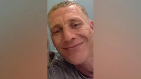 Humberside Police Shane Boxall has short-cropped blond hair and hazel eyes. He is wearing a grey T-shirt and is smiling into the camera.