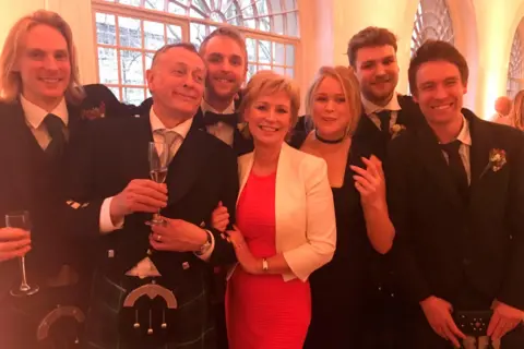 Sally Magnusson Sally Magnusson with her family in formal wedding clothes. Sally is in the centre wearing a red dress and white jacket and her daughter Anna Lisa is on her right wearing a black dress. Sally's husband Norman is on her left in a kilt and sporran and formal wedding jacket. Also pictured are the couple's four sons Jamie, Rossie, Magnus & Siggy.