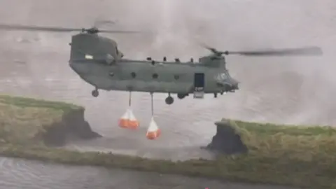 Large helicopter drops off heavy bags to stop flooding that has created a huge hole in a grassy bank.