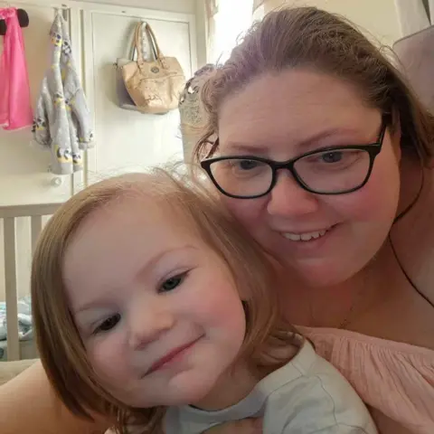 Kelly Ann Bigham Kelly Ann Bigham, in a peach top and glasses, hugs her 22-month old daughter Alana, a smiling light brown-haired girl , wearing a white t-shirt.