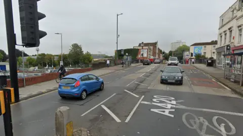 Picture of Lawrence Hill area. It's a street with vehicles headiing in different directions with cream coloured buildings in the background. There is a cyclist riding on the pavement. 