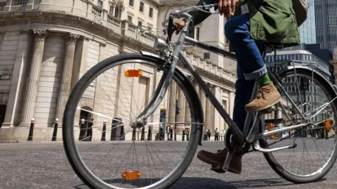 A man in blue jeans, a green coat and brown shoes on a bicycle in a city