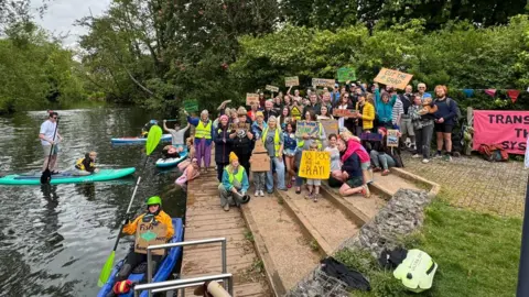 Jo MacDonald People are sat down on a set of steps leading down to a river, holding many different home-made placards with slogans against sewage