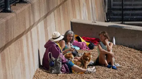 Three women sit on pebble beach in Dover. They are in summer clotheses. One is wearing a sun hat, one is in a swimming top, with the third in shorts.
