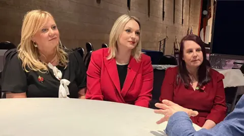 Nurses Lisa Lockey, Bethany Hutchison and Karen Danson sit at a table facing a reporter asking them questions. Lisa and Bethany both have long blonde hair and Karen has dark red hair. They are all smartly dressed with two of them displaying poppies.

