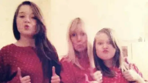 Family photo Two young girls with long brown hair, with a woman with long blonde hair in between them. They are all wearing matching red pyjamas, pulling pouting faces and have their thumbs up as they look at the camera.