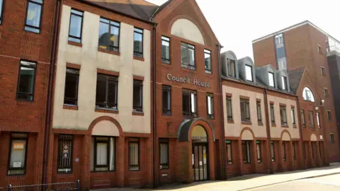 A red brick and white frontage of a modern building with three floors and many windowns with a large silver sign on the front of the building saying council house 