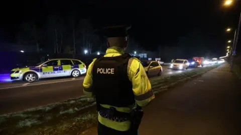 PA Media A police officer wearing a flak jacket with police written across the back. They're also wearing a bright yellow jacket underneath with police branding on it. A police car can be seen in the background.